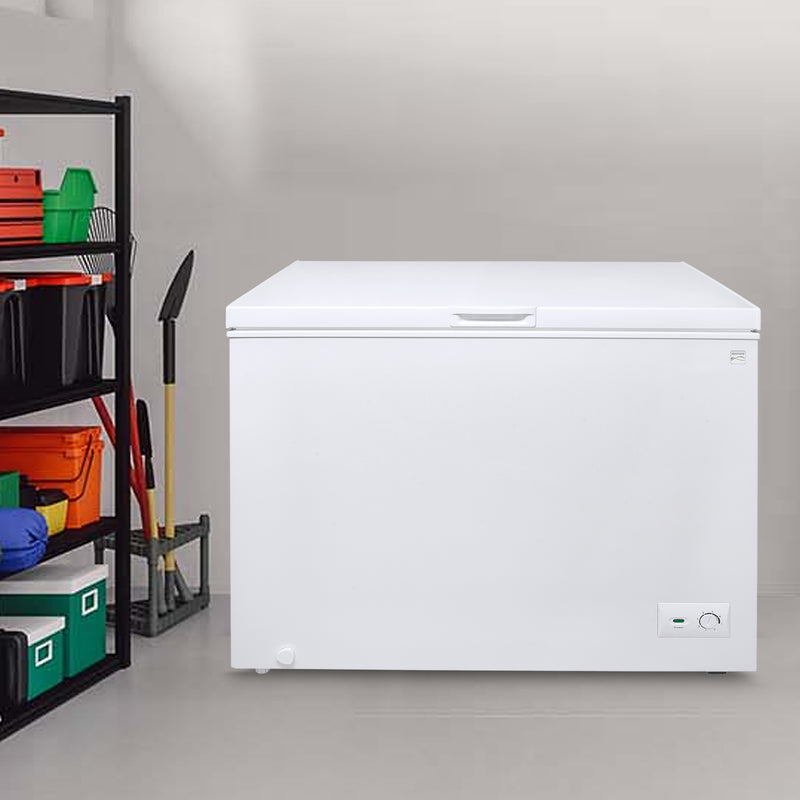 Kenmore chest freezer, closed, with a light gray wall behind it and a utility shelf with camping gear, storage totes, and tools to the left