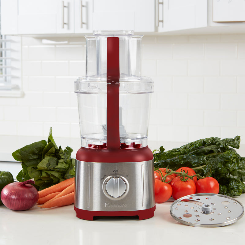 Kenmore 11 cup food processor and vegetable chopper, empty, on a light gray countertop with white tile backsplash behind. There is a bowl of chopped tomatoes to the left and a cutting board, knife, and whole and chopped ingredients to the right.