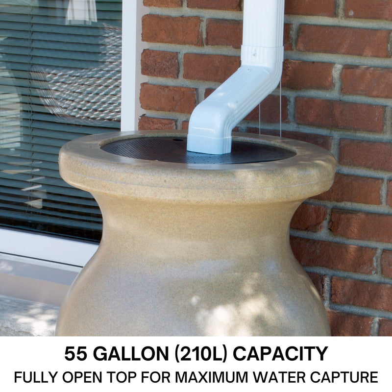 Closeup of the top half of a Koolscapes decorative sandstone-look rainwater urn set up below a white downspout with a red brick wall in the background. Text below reads, "55 gallon (210L) capacity: Fully open top for maximum water capture."