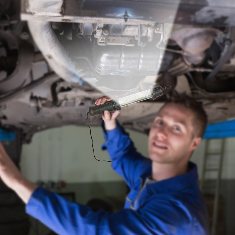 Lifestyle image of a person with light skin and crew cut brown hair wearing a bright blue coverall standing under a vehicle hoist and holding the 12V work light as a flashlight to illuminate the underside of a vehicle