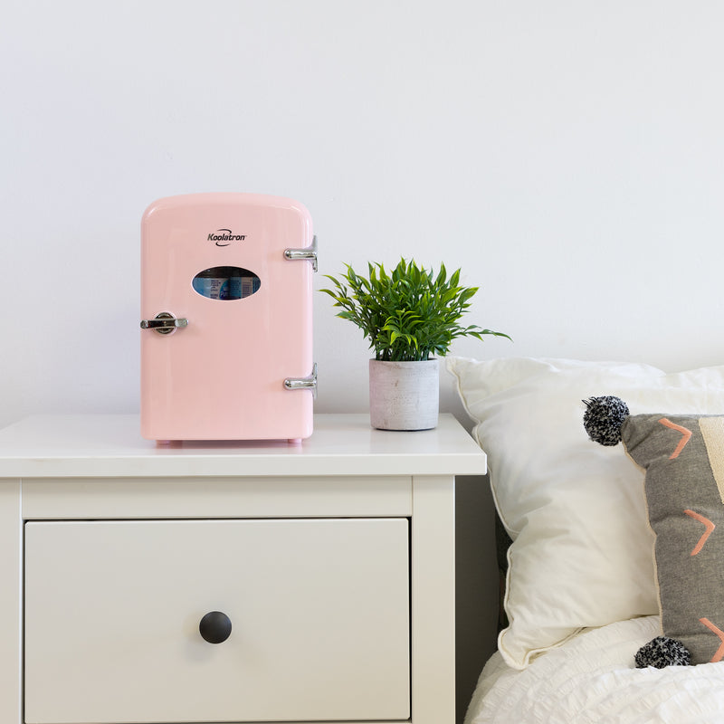 Lifestyle image of Koolatron retro 6 can mini fridge, closed, with a plant in a white pot on its right, on a white bedside table beside a bed with white linens 